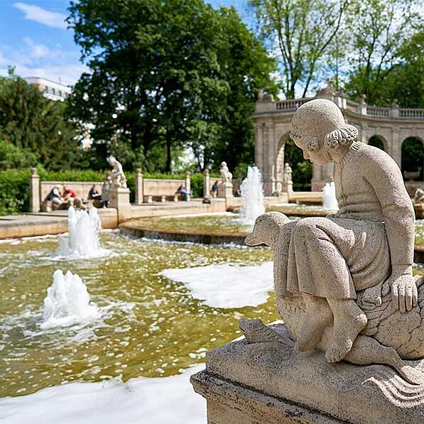 Im Detail zeigt das Bild einen großen, rechteckigen Springbrunnen mit mehreren Springbrunnen, die Wasser in die Luft spritzen. Das Wasser erscheint leicht trüb und schäumt um die Basis der Springbrunnen herum. Um den Springbrunnen herum befinden sich mehrere Steinbänke, auf denen sich Menschen entspannen. Im Hintergrund steht eine steinernen Architektur, die einem Pavillon ähnelt, mit Bögen und Säulen. Mehrere Bäume mit dichtem, grünem Laub umgeben den Springbrunnen und den Pavillon. Die Gesamtatmosphäre ist friedlich und ruhig.
Im Vordergrund sitzt eine detaillierte Steinstatue eines jungen Mädchens, das ein langes Kleid trägt und auf einem Tier sitzt, das wie ein Vogel oder ein kleines Tier aussieht. Die Statue ist aus hellem Stein gefertigt und zeigt feine Details in Kleidung und Gesichtszügen des Mädchens. Die Statue strahlt Ruhe und Beschaulichkeit aus.
Die Farben des Bildes sind natürlich und gedämpft, mit Grüntönen der Bäume, den helleren Farbtönen des Steins und dem braunen bis grünlichen Wasser des Brunnens. Das Licht scheint weich und diffus zu sein, was auf einen sonnigen, aber nicht grellen Tag hindeutet. Die gesamte Szene vermittelt ein Gefühl von Frieden, Ruhe und Schönheit.