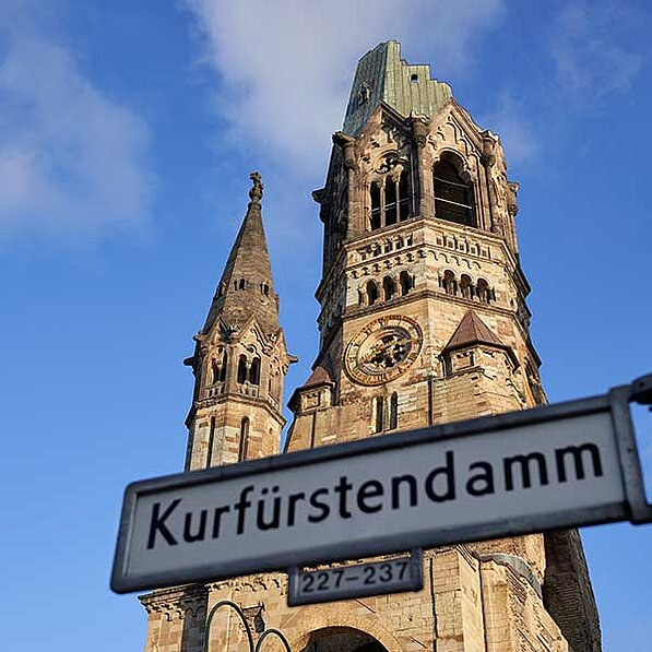 Das Bild zeigt einen niedrigen Blickwinkel auf die Gedächtniskirche in Berlin, Deutschland. Im Vordergrund befindet sich ein Straßenschild mit der Aufschrift „Kurfürstendamm 227-237“. Die Kirche ist aus Sandstein und besteht aus zwei Türmen. Der höhere Turm ist teilweise zerstört, mit einem fehlenden oberen Teil und sichtbaren Schäden. Der kleinere Turm ist intakt und zeigt eine spitz zulaufende Spitze. Die Uhr des höheren Turms ist sichtbar und scheint intakt zu sein. Der Himmel ist hellblau mit einigen wenigen Wolken. Die Atmosphäre ist ruhig und nachdenklich, wobei der Blick auf die beschädigte Kirche an die Geschichte und die Zerstörung erinnert. Die Stimmung ist eine Mischung aus Trauer und Hoffnung, da die teilweise zerstörte Kirche als Mahnmal steht, gleichzeitig aber auch die Wiederauferstehung und den Wiederaufbau symbolisiert.