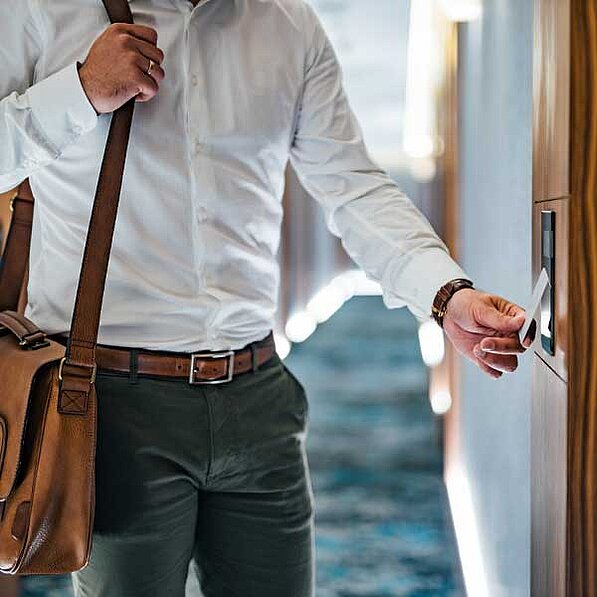 Im Detail sieht man einen Mann, der einen weißen Hemd und eine dunkelgrüne Hose trägt. Er hat einen braunen Ledergürtel und eine braune Ledertasche über der Schulter. Er hält eine Schlüsselkarte in der Hand und hält sie an einen elektronischen Türöffner an einer Holztür in einem Hotelflur. Die Atmosphäre ist ruhig und professionell. Der Flur ist hell und modern eingerichtet. Der Mann wirkt selbstbewusst und effizient. Die allgemeine Stimmung ist neutral, aber die Szene vermittelt ein Gefühl von Reise und Geschäft.