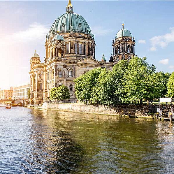 Im Detail zeigt das Bild die Berliner Dom, ein imposantes Gebäude aus Sandstein mit einer großen, grün kupfernen Kuppel. Die Architektur ist im barocken Stil gehalten, mit vielen Details und Ornamenten. Zweierlei Türme flankieren die Hauptkuppel. Vor dem Dom erstreckt sich eine Reihe von grünen, üppigen Bäumen entlang eines steinernen Ufers, das den Fluss vom Land trennt. Der Fluss selbst ist ruhig und reflektiert das Licht des sonnigen Himmels. Im Hintergrund ist ein Teil von weiteren Gebäuden der Stadt zu erkennen, sowie eine Brücke, über die ein kleines Boot fährt. Die Sonne scheint von der linken Seite des Bildes, wodurch ein sanftes, warmes Licht auf die Szene fällt. Die Gesamtatmosphäre ist friedlich, ruhig und malerisch, und vermittelt ein Gefühl von Schönheit und Großartigkeit. Das Bild strahlt eine positive und ruhige Stimmung aus, die den Betrachter einlädt, die Schönheit des Ortes zu genießen.