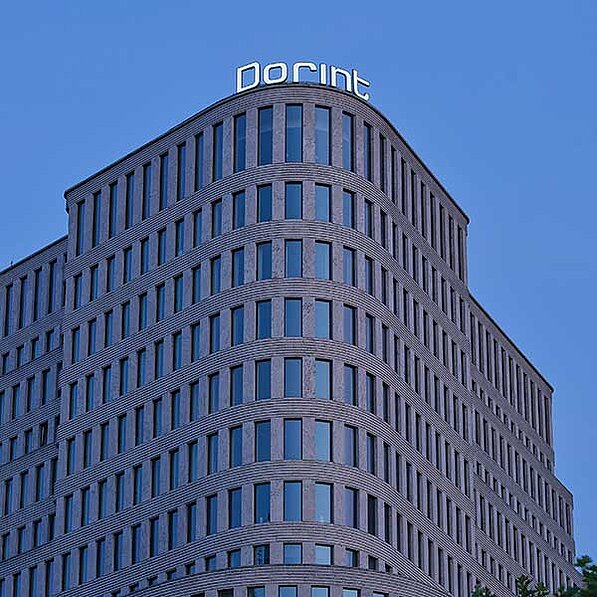 Im Detail zeigt das Bild die Fassade eines mehrstöckigen Gebäudes. Die Fassade ist durchzogen von vertikalen Fensterreihen, die in einem regelmäßigen Muster angeordnet sind. Zwischen den Fensterreihen befinden sich graue oder beige Säulen, die das Gebäude strukturieren. Das Gebäude scheint eine leicht abgerundete Ecke zu haben. Auf der Spitze des Gebäudes ist das Wort "dorint" in leuchtenden Buchstaben zu sehen. Der Himmel im Hintergrund ist klar und blau, was eine ruhige und friedliche Atmosphäre vermittelt. Im unteren rechten Bereich des Bildes sind einige grüne Baumkronen erkennbar, die darauf hindeuten, dass sich das Gebäude in einer urbanen Umgebung mit etwas Baumbestand befindet. Die Stimmung des Bildes ist sachlich und architektonisch, wobei der Fokus auf der klaren, modernen Linienführung des Gebäudes liegt.