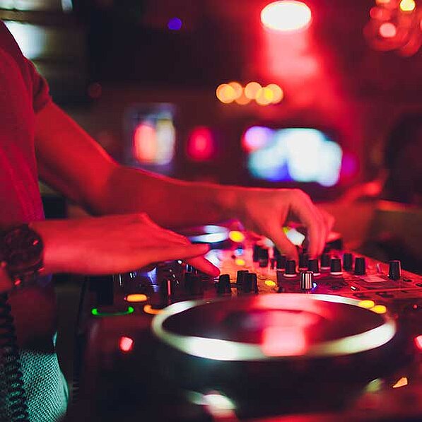 Das Bild zeigt die Hände eines DJs, die über einem Mischpult in einem schwach beleuchteten Raum schweben. Die Beleuchtung ist überwiegend rot und erzeugt eine warme, aber auch etwas düstere Atmosphäre. Die Hände des DJs sind im Vordergrund scharfgestellt, während der Hintergrund unscharf ist und nur die Umrisse weiterer Objekte zeigt, wie z.B. Lichter, die an eine Bar oder einen Club erinnern. Die roten und orangefarbenen Lichter im Hintergrund verstärken die Stimmung eines nächtlichen, pulsierenden Ortes. Der DJ trägt eine Uhr am Handgelenk. Die Oberflächen des Mischpults glänzen leicht und reflektieren das rote Licht. Das Bild konzentriert sich auf den Akt des Mixens und die damit verbundenen Bewegungen an den Reglern und Knöpfen des Mischpults – es ist ein dynamischer und intensiver Moment eingefangen. Die Gesamtstimmung ist pulsierend, energiegeladen und etwas mysteriös aufgrund der Dunkelheit und des Fokus auf die Hände.