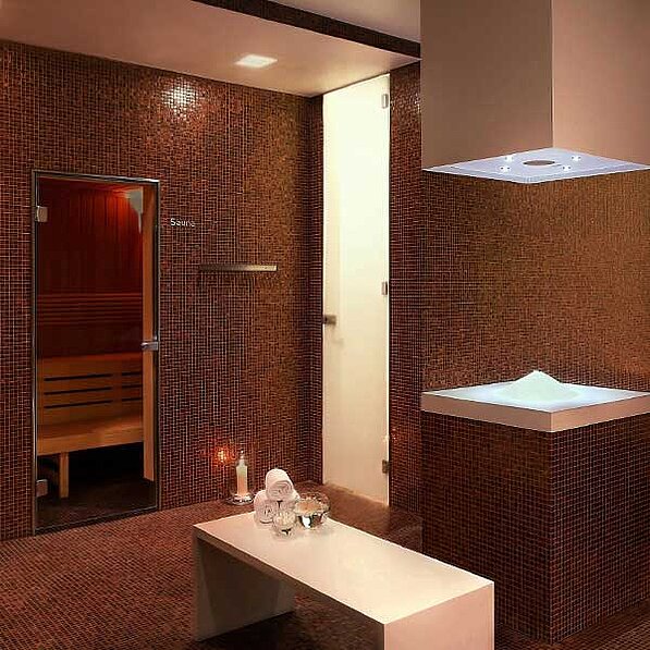 Im Detail zeigt das Bild einen Raum, der mit dunkelroten Mosaikfliesen verkleidet ist. Im Vordergrund steht eine weiße Bank. Auf der Bank stehen zwei weiße Handtücher, zwei brennende Kerzen und eine Glasschale mit weißem Sand oder Salz. Rechts befindet sich ein weißer, quadratischer Behälter, der mit weißem Sand oder Salz gefüllt ist und auf einem Podest aus den gleichen Mosaikfliesen steht wie die Wände. Links im Raum ist eine verglaste Sauna mit Holzinnenausstattung zu sehen, die den Raum in zwei Bereiche teilt. Über dem weißen Behälter ist eine quadratische, eingebaute Deckenleuchte angebracht. Die gesamte Atmosphäre ist ruhig, entspannend und luxuriös. Die Beleuchtung ist gedämpft und warm. Es sind keine Personen oder Tiere auf dem Bild zu sehen.