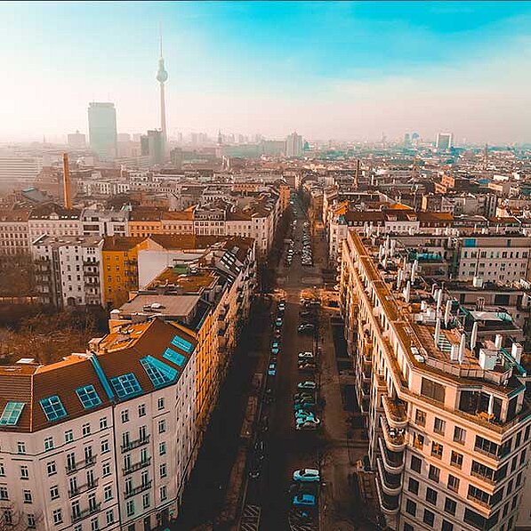 Das Bild zeigt eine atemberaubende Luftaufnahme eines Teils von Berlin an einem sonnigen Tag. Im Vordergrund sind mehrere mehrstöckige Gebäude mit roten Ziegeldächern und hellen Fassaden zu sehen. Die Gebäude stehen eng aneinander und scheinen Teil einer Wohngegend zu sein. Eine Straße schlängelt sich durch die Bebauung, auf der zahlreiche Autos geparkt sind. Die Straße ist relativ schmal und scheint nicht viel Verkehr zu haben.
Im mittleren und Hintergrund erstreckt sich die Stadt Berlin bis zum Horizont. Viele weitere Gebäude, in verschiedenen Größen und Architekturstilen, sind zu erkennen. Es ist eine dichte Bebauung, die auf eine urbane Umgebung hinweist. In der Ferne ist der Berliner Fernsehturm deutlich sichtbar, was die Perspektive und den Umfang der Stadt unterstreicht.
Die Atmosphäre des Bildes ist ruhig und friedlich, trotz der urbanen Umgebung. Die warme Farbgebung der Gebäude und der leicht neblige Himmel erzeugen eine warme und gemütliche Stimmung. Das Bild vermittelt ein Gefühl von Ordnung und Struktur, gleichzeitig aber auch von Großstadt-Charakter. Die Perspektive von oben erlaubt einen umfassenden Überblick und betont gleichzeitig die Details der Architektur und das Stadtbild. Die Farbgebung ist leicht gesättigt, mit warmen Tönen, die den Eindruck von Sonnenlicht und Wärme verstärken.