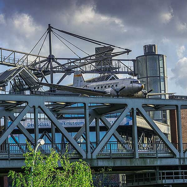 Das Bild zeigt eine Douglas C-47 Skytrain, ein zweimotoriges Transportflugzeug, das auf einer modernen Stahlbrücke ruht. Das Flugzeug ist silbern-grau und trägt die Aufschrift "European Air Transport Service". Es wirkt etwas verwittert, was auf sein Alter und seine Geschichte hinweist. Die Brücke ist eine stählerne Konstruktion mit vielen diagonalen und vertikalen Trägern, die eine robuste und industrielle Ästhetik vermitteln. Das Flugzeug liegt auf einem Teil der Brücke, der sich über den Eingang eines Gebäudes erstreckt. Dieses Gebäude hat eine moderne Glasfassade und einen zylindrischen Turm aus Glasbausteinen. Auf der Fassade ist teilweise die Aufschrift "Deutsches Technikmuseum" zu erkennen, was darauf schließen lässt, dass sich das Flugzeug in einem Museum befindet.
Die Atmosphäre des Bildes ist eine Mischung aus Industrie und Geschichte. Der graue Himmel und die stählerne Struktur der Brücke verleihen dem Bild einen etwas melancholischen und zugleich imposanten Charakter. Das Flugzeug als zentrales Element steht für vergangene Zeiten und technisches Können. Der Kontrast zwischen dem alten Flugzeug und der modernen Architektur des Museums unterstreicht diesen Aspekt. Im Vordergrund ist ein Teil eines grünen Baumes zu sehen, der eine Kontrastfarbe zum Grau der Brücke und des Flugzeugs bietet und einen Hauch von natürlicher Umgebung hinzufügt.