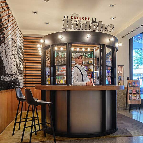 Das Bild zeigt einen kreisförmigen Kiosk, der sich in einem hellen, modern gestalteten Innenraum befindet. Der Kiosk ist mit "Et Kölsche Büdche" beschriftet, was auf einen Kiosk mit regionalen Kölner Produkten hindeuten könnte. Ein junger Mann mit Schiebermütze und Hosenträgern steht hinter der Theke, was den Eindruck eines Verkäufers oder Betreibers vermittelt.
Die Theke ist aus Holz und hat eine graue, matte Front. Hinter dem Mann sind Glasscheiben zu sehen, die Einblick in das Innere des Kiosks geben, wo verschiedene Produkte wie Getränke und Snacks in Regalen aufgereiht sind. Die Oberseite des Kiosks ist mit Glühbirnen umrandet, die für eine warme, einladende Beleuchtung sorgen.
Links neben dem Kiosk stehen zwei schwarze Barhocker, die zum Verweilen einladen. An der Wand im Hintergrund ist ein abstraktes Kunstwerk zu sehen, das aus vielen kleinen, wellenförmigen Elementen besteht und ein Gesicht zu formen scheint. Die Wandverkleidung besteht aus horizontalen Holzpaneelen, was dem Raum eine natürliche Note verleiht.
Rechts vom Kiosk befindet sich ein großes Fenster, durch das Tageslicht einfällt. Davor steht ein Display mit verschiedenen kleinen Produkten oder Souvenirs. Der Boden ist mit einem Teppich oder einer Matte in einem dezenten Muster bedeckt.
Die Atmosphäre ist einladend und modern, mit einem Hauch von Retro-Charme durch die Kleidung des Mannes und das Design des Kiosks. Die Beleuchtung und die Farbgebung sind warm und freundlich, was den Raum gemütlich wirken lässt.