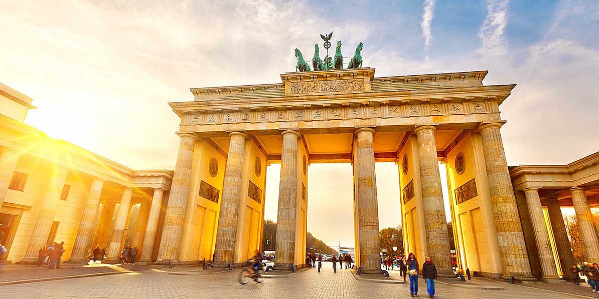 Das Bild zeigt das Brandenburger Tor in Berlin im goldenen Abendlicht, umgeben von Menschen, die zu Fuß gehen und Fahrrad fahren.